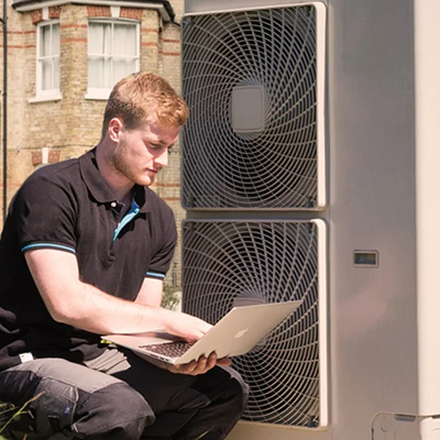 Heating engineer using a laptop to configure and commission an outdoor air source heat pump unit against a brick wall.